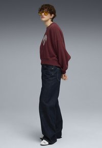 Mørkerød oversized sweatshirt med grafisk detalje; mørkeblå vide bukser; hvide sko; gule solbriller; kort krøllet hår.