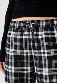 Persona vestida con pantalones de pijama de cuadro en blanco y negro con cordón y una camiseta negra de manga larga que muestra el abdomen.