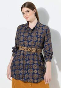 Camicia blu a fantasia con dettagli dorati, maniche lunghe e chiusura a bottoni, abbinata a una cintura intrecciata testurizzata con fibbia circolare.