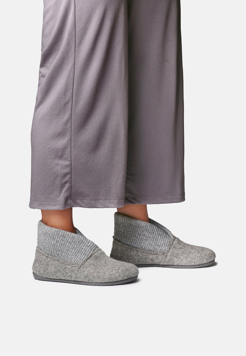 Personne portant un pantalon gris ample et des chaussons d'intérieur gris doux avec de hauts poignets côtelés sur un fond uni.