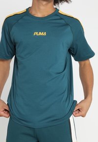 Teal idrottströja med gula detaljer på axlarna, prydd med Puma-logotypen i gult. Lättviktsmaterial och avslappnad passform.