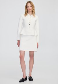 Chaqueta peplum blanca con cuello y seis botones metálicos, combinada con una falda blanca a juego. Llevada con mocasines negros. Textura suave.