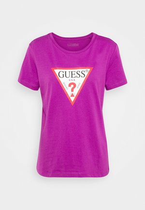 Purppura puuvillainen t-paita, jossa on kolmiomainen graafinen logo valkoisena ja punaisena, tekstit "GUESS" ja "U.S.A." Lyhyet hihat ja pyöreä kaula-aukko.