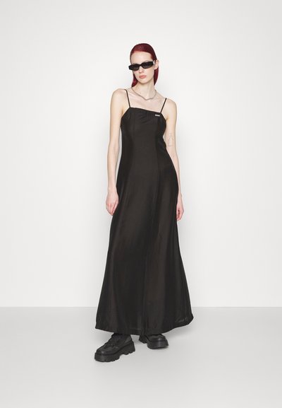 Čierna saténová maxi šaty s tenkými ramienkami, štvorcovým výstrihom a voľnou siluetou. Modelka má na sebe masívne čierne topánky a slnečné okuliare.