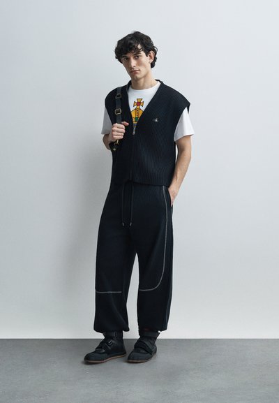 Μαύρο ριπ γιλέκο πάνω από λευκό T-shirt με γραφικό, σε συνδυασμό με φαρδιά μαύρα παντελόνια με λευκό ράψιμο και μαύρα αθλητικά παπούτσια με λουράκια.