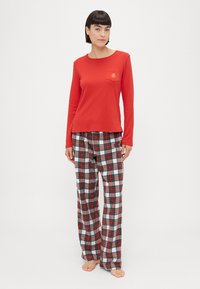 Rdeča majica s dolgimi rokavi in majhno žepno logotip, v kombinaciji s karirastimi pižam pants v rdečih, belih in modrih odtenkih. Mehka tkanina in udoben kroj.