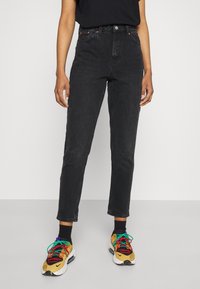 Persona che indossa jeans neri a vita alta, calze nere e sneaker gialle e nere con lacci multicolori, in piedi su un pavimento grigio.