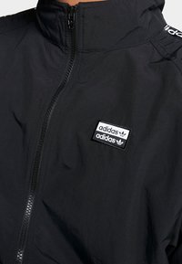 Svart zip-up jacka med hög krage, prydd med en broderad rektangulär Adidas-logotyp på bröstet, tillverkad av lätt material.