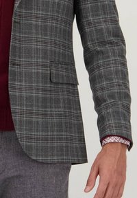 Grijze geruite blazer met gestructureerde schouders, een enkele zak en contrasterende bordeauxrode manchetten. Getextureerde stof met een subtiele glans en fijne details.