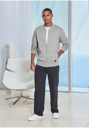 Mann trägt graue Reißverschlussjacke, weißes Hemd, schwarze Hose und weiße Turnschuhe, steht drinnen in der Nähe eines weißen Stuhls und eines Glasfensters.