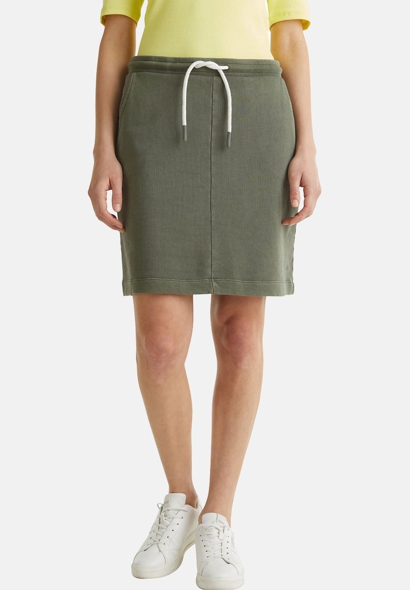 Femme portant une jupe olive à cordon, un top jaune et des baskets blanches, debout devant un fond blanc uni.