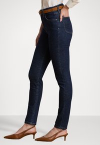 Donkerblauwe skinny jeans met een gladden denimtextuur, voorzien van een middelhoge tailleband en een hand in een zak gestoken. Gekocht met bruine accenten.