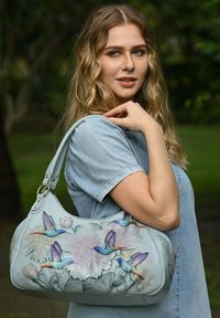 Lichtblauwe leren handtas met bloem- en kolibrid ontwerpen, met een gebogen vorm en een schouderband versierd met metalen accenten.