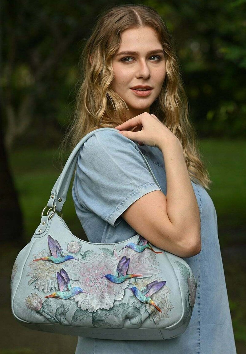 Lichtblauwe leren handtas met bloem- en kolibrid ontwerpen, met een gebogen vorm en een schouderband versierd met metalen accenten.