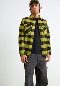Sort og gulternet flannel skjorte med to brystlommer, knaplukning, båret over en sort t-shirt, parret med mørkegrå jeans.