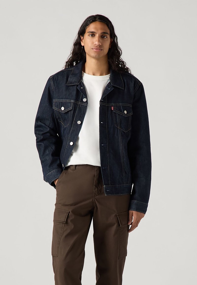 Donker denim jack met een klassiek ontwerp, twee borstzakken en zilverkleurige accenten. Gecombineerd met een wit shirt en bruine cargobroek.