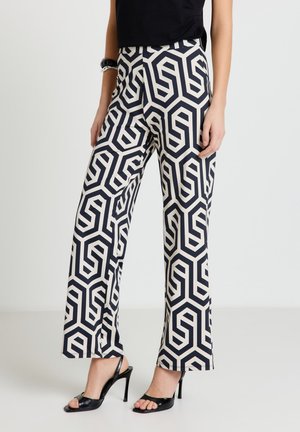 Donna che indossa pantaloni a gamba larga con motivo geometrico bianco e nero, sandali con tacco neri e una maglia nera, in piedi contro uno sfondo semplice.