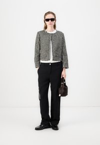 Jachetă cropped în alb și negru cu model houndstooth, cămașă albă, pantaloni negri cu buzunare cargo, pantofi negri și o mică poșetă maro.