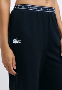 Zwarte joggingbroek met een gladde structuur, elastische tailleband met het merk "LACOSTE" en een klein wit krokodillenlogo op de dij.