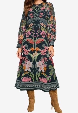 Nainen, joka on pukeutunut pitkähihainen, tummakuvioinen midimekko ja ruskeat mokkanahasta tehdyt polven yli yltävät saappaat, seisomassa yksiväristä taustaa vasten.