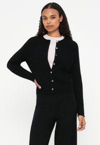 Cardigã preto de malha com decote arredondado, fecho frontal com botões e mangas compridas, usado sobre uma blusa rosa clara. Textura suave.