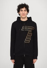 Black zip-up hoodie in soft material featuring a gold striped graphic design on the chest. Includes a hood and front pockets.
