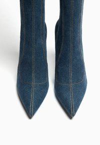 Džínové kotníkové boty s špičatými prsty, vyrobené z modré textilie, s kontrastním žlutým prošíváním a přiléhavým střihem.