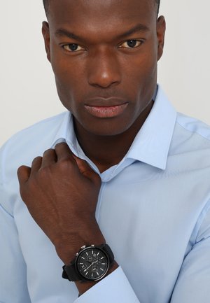 Black wristwatch with a textured strap and round dial, featuring silver accents and multiple subdials, worn on a wrist with a buttoned blue shirt.