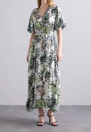 Femme portant une longue robe à imprimé floral avec des manches bouffantes et des sandales noires à talons hauts et lanières, debout sur un sol gris.