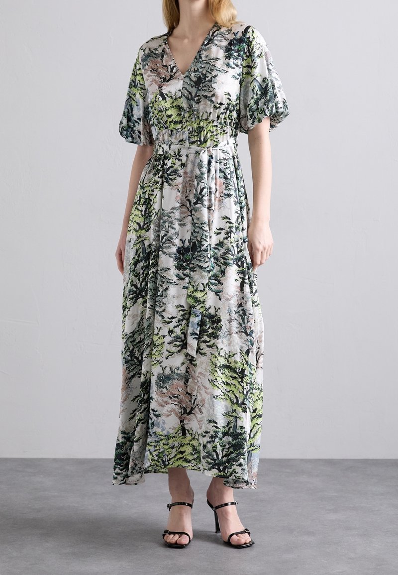 Femme portant une longue robe à imprimé floral avec des manches bouffantes et des sandales noires à talons hauts et lanières, debout sur un sol gris.