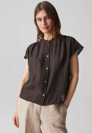 Vrouw die een zwarte blouse met korte mouwen en witte krijtstrepen draagt, en beige broek, staat met één hand in de zak.