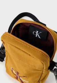 Mała torebka na ramię z żółtego nylonu w kolorze musztardowym z bordowym wnętrzem. Posiada zamek błyskawiczny i metkę z logo wewnątrz. Teksturowana powierzchnia z przeszyciami.