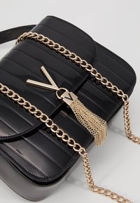 Borsa quadrata in pelle verniciata nera con trama a coste orizzontali, tracolla con catena dorata e decorazione con nappina in metallo.