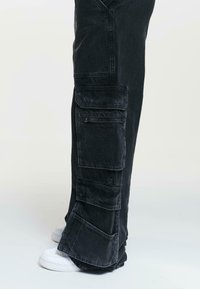 Black cargo pants made of denim, featuring multiple large pockets and a relaxed fit. Worn with white sneakers.