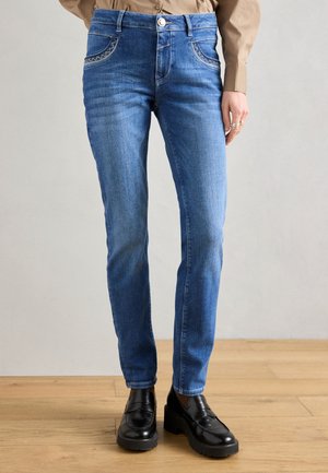 Μπλε τζιν παντελόνι με εφαρμοστή γραμμή, διακοσμητικές ραφές στις μπροστινές τσέπες και κλείσιμο με ένα κουμπί. Φορεμένο με μαύρα μοκασίνια.