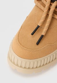 Punta della scarpa in suede marrone con superficie texturizzata, caratterizzata da lacci arancioni cuciti e punte dei lacci nere, e una suola beige spessa.
