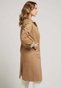 Manteau trench beige avec un col, des poches latérales et une doublure à motif écossais. Il présente une fente à l'arrière pour plus de mobilité.