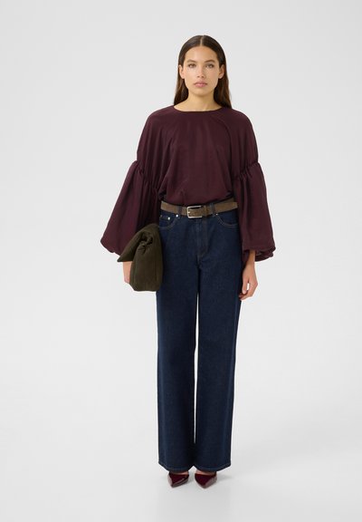 Haut bordeaux à manches bouffantes avec un col rond, associé à un jean large en denim foncé taille haute et un sac vert. Des chaussures à talons complètent le look.
