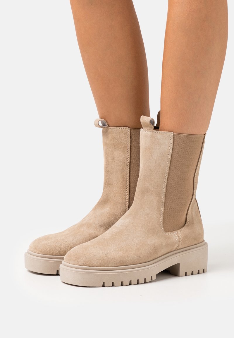 Bottes cheville en daim beige avec semelles en caoutchouc épaisses et panneaux élastiques sur les côtés, portées sur des jambes nues sur fond blanc.