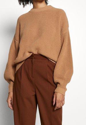 Personne portant un pull en maille côtelée beige avec des manches bouffantes, rentré dans un pantalon large marron taille haute.