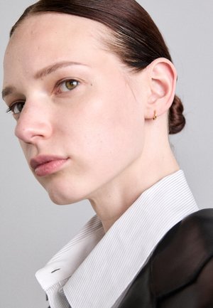 Pendiente de aro dorado, piel clara, cabello castaño claro recogido en un moño, vistiendo una camisa negra con un cuello a rayas blancas, perfil lateral.