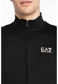 Giacca nera con zip realizzata in tessuto liscio e con colletto alto. Presenta il logo EA7 Emporio Armani sul lato sinistro del petto in bianco e oro.