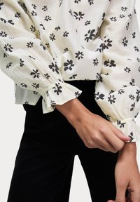 Blusa bianca con stampa floreale nera, texture plissettata, maniche lunghe e polsini arricciati. Abbinata a pantaloni neri.