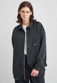 Νέος άνθρωπος φοράει ένα υπερμεγέθες μαύρο πουκάμισο με κουμπιά πάνω από λευκή μπλούζα και μαύρο παντελόνι, στέκεται σε έναν απλό ανοιχτό γκρι φόντο.