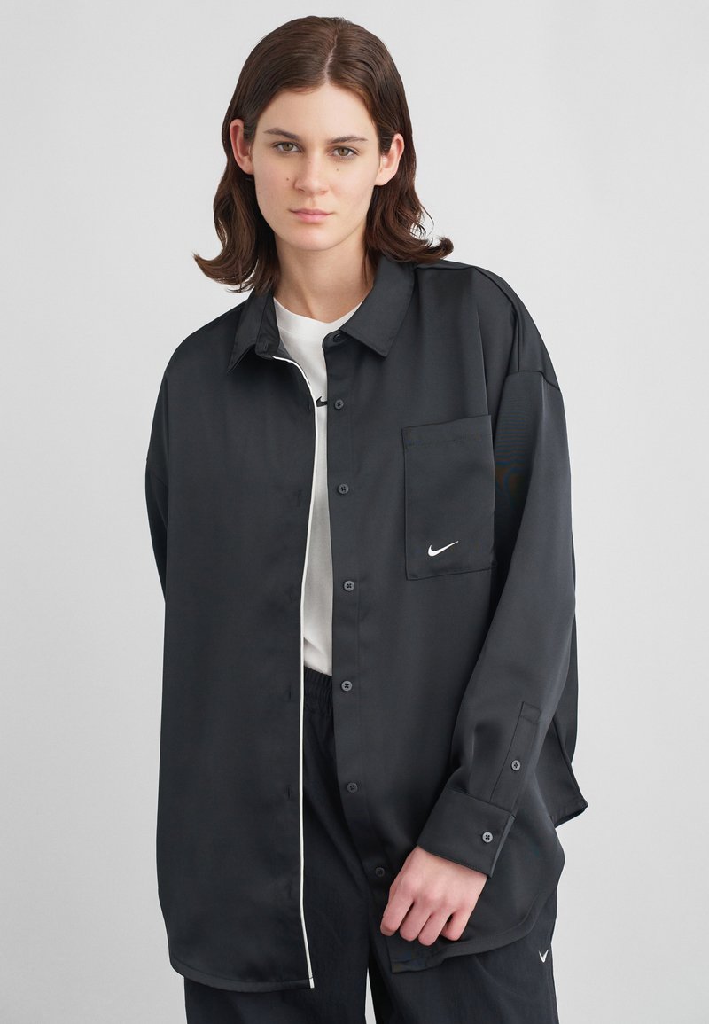 Νέος άνθρωπος φοράει ένα υπερμεγέθες μαύρο πουκάμισο με κουμπιά πάνω από λευκή μπλούζα και μαύρο παντελόνι, στέκεται σε έναν απλό ανοιχτό γκρι φόντο.