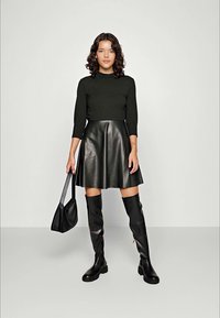 Haut noir à manches longues associé à une jupe en cuir noir évasée. Le mannequin porte des bottes noires jusqu'aux genoux et porte un petit sac à bandoulière noir.