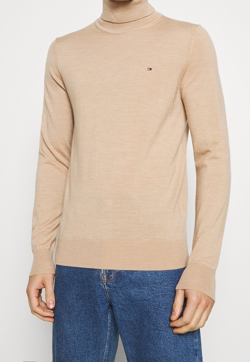 Pull à col roulé beige en tissu doux, avec une coupe ajustée et des poignets côtelés. Petit logo sur la poitrine. Associé à un jean bleu.