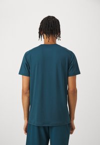 Tyrkysová košile s krátkým rukávem vyrobená z měkké látky, s kulatým výstřihem a minimem švů, viděná ze zadní strany.