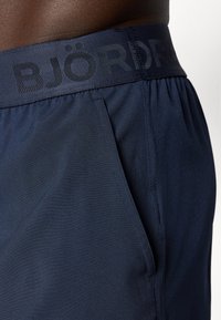 Bermudas azul-marinho feitas de tecido leve, com uma cintura elástica com "BJÖRD" impresso em cinza-escuro. Bolsos laterais visíveis.