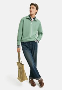 Hellgrüner Fleece-Pullover mit Viertel-Reißverschluss, über ein kariertes Hemd getragen. Kombiniert mit dunkelblauen Jeans, braunen Schuhen und einer sandfarbenen Tragetasche. Einfaches Design und Materialien.